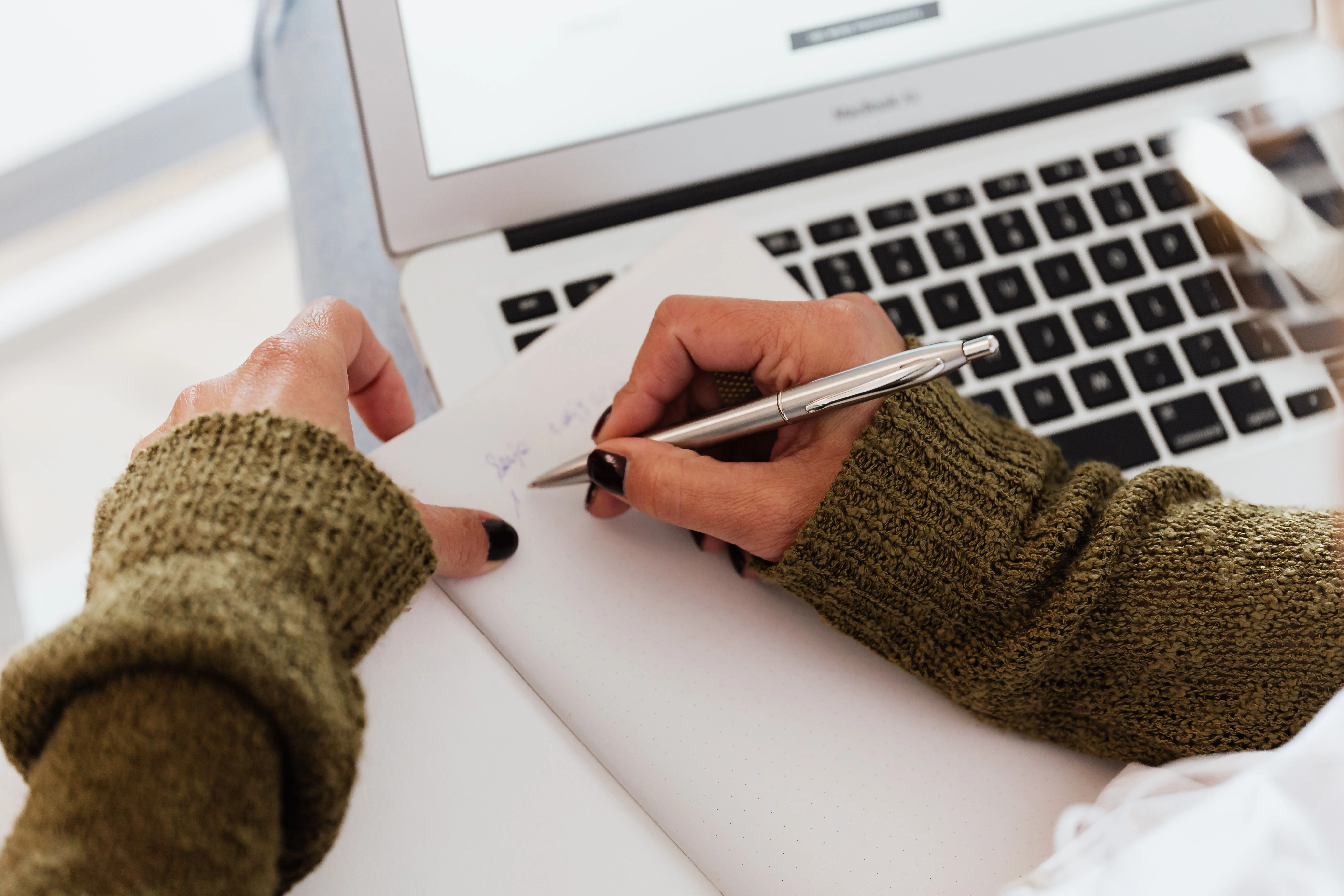 Close-up of a young woman writing notes on a notepad with a laptop, indicating a study or work setting.